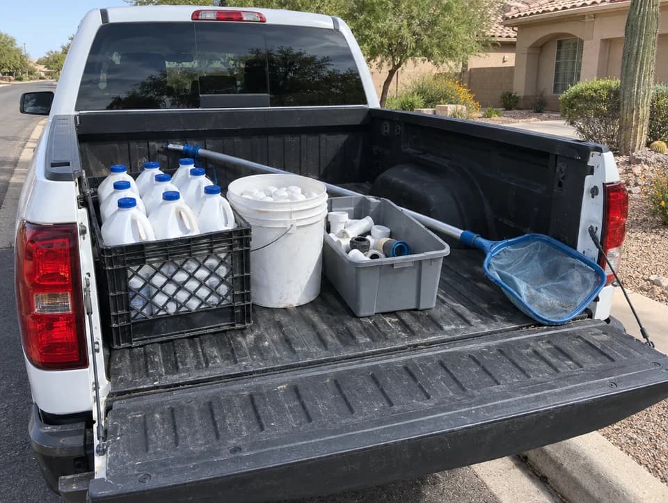 The open bed of a white pool service pickup at the curb, loaded with a crate of liquid chlorine jugs, a bucket of chlorine tablets, a grey bin of plumbing fittings and a leaf net